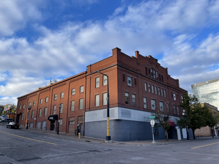 Exterior view of a three-story brick building positioned on a downtown street corner. A tall new glass-enclosed building is visible in the background to the right.
