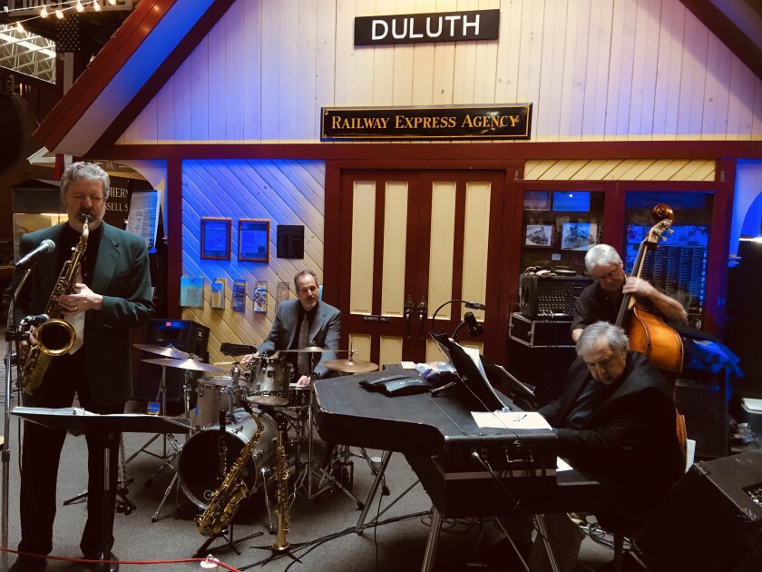 Four musicians play jazz music in front of a yellow-and-red model train depot. From left, they play saxophone, drum, keyboard and upright bass.
