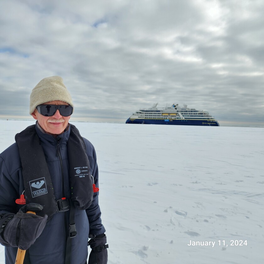 A man bundled up for the cold is pictured with hat and dark sunglasses with a large cruise ship in the background