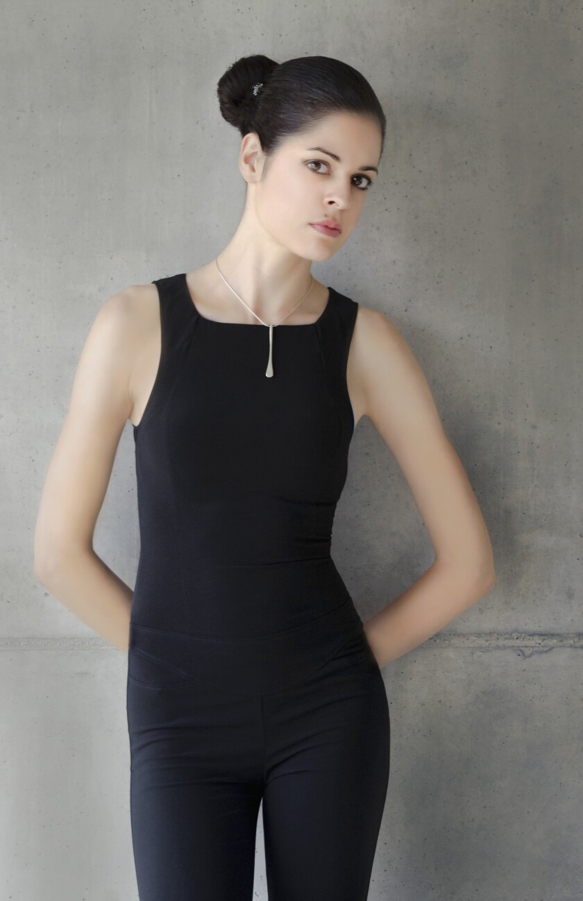 Woman in form-fitting black clothing stands with her hands behind her back, leaning against a concrete wall and looking severe.