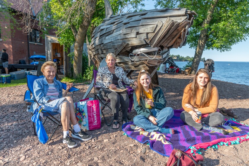 Four light-skinned people of varying ages sit smiling on a rocky lake beach in front of a large wooden shark sculpture.