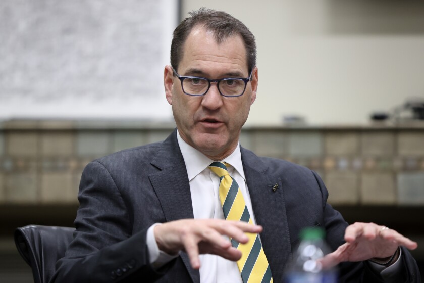 A man in a charcoal suit, white shirt and green and yellow tie gestures with both hands.