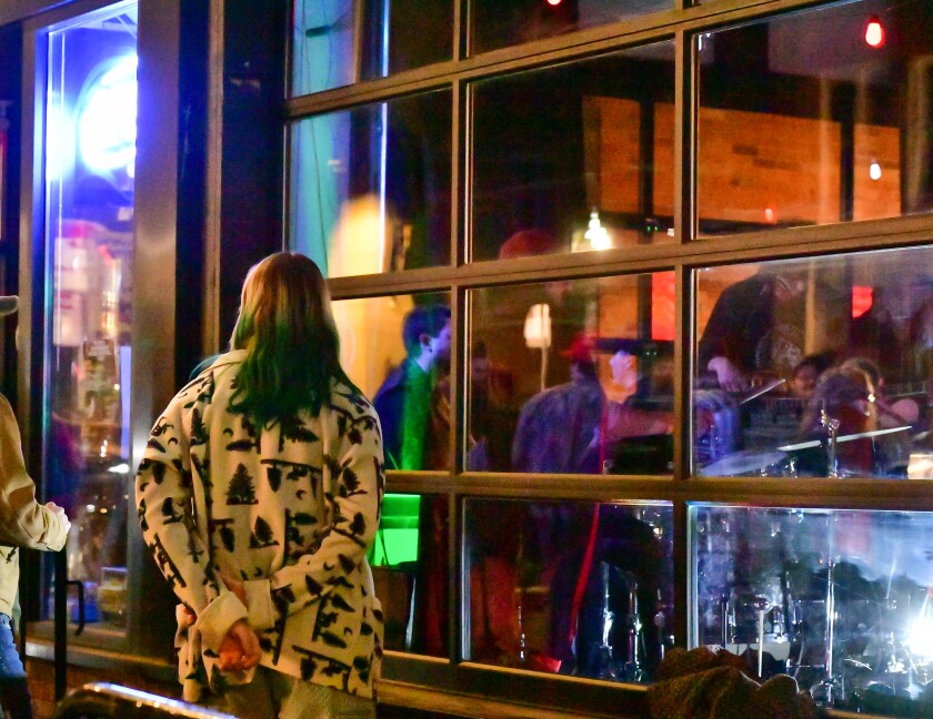 Pessoa com longos cabelos verdes fica do lado de fora da janela de um bar