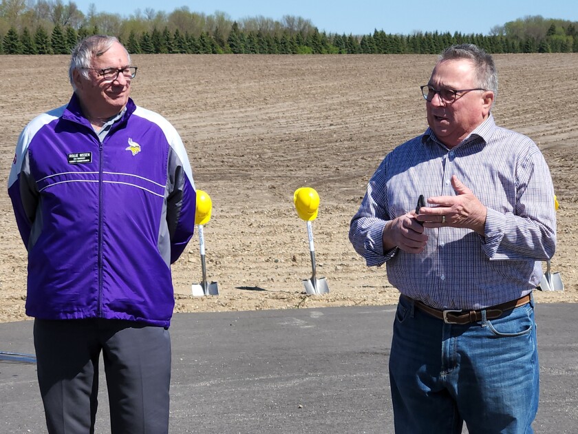 Two men standing in front of a dirt covered field and shovels topped in yellow hard hats speaking to attendees of a groundbreaking ceremony