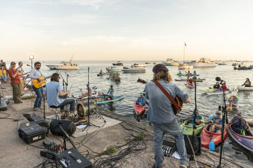 Band of light-skinned men performs on a concrete pier on a large lake, with spectators positioned in kayaks and motorboats.