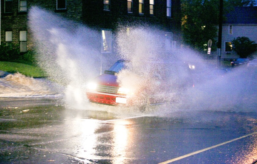 A red minivan drives through flooded streets, creating a large splash on either side