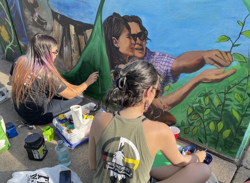 Two people paint a mural of individuals examining leaves in a farm-like setting.