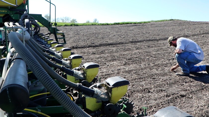 A farmer kneels to check soybean seed depth behind his John Deere planterat left.