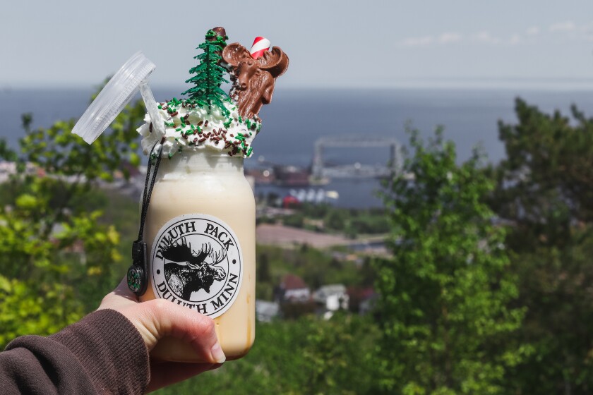 A hand holds a jar full of a brown-tan malt mixture, bearing a Duluth Pack sticker and topped with copious whipped cream as well as a small tree and a chocolate moose, with the Aerial Lift Bridge and Duluth Harbor visible in the background.