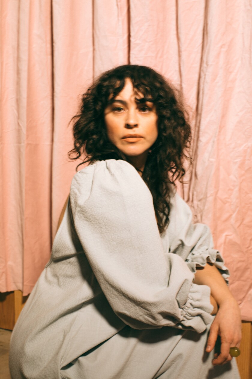 A woman in a baggy light gray dress sits in front of peach colored curtains and turns around to look at the camera. Her hair is black and curly.