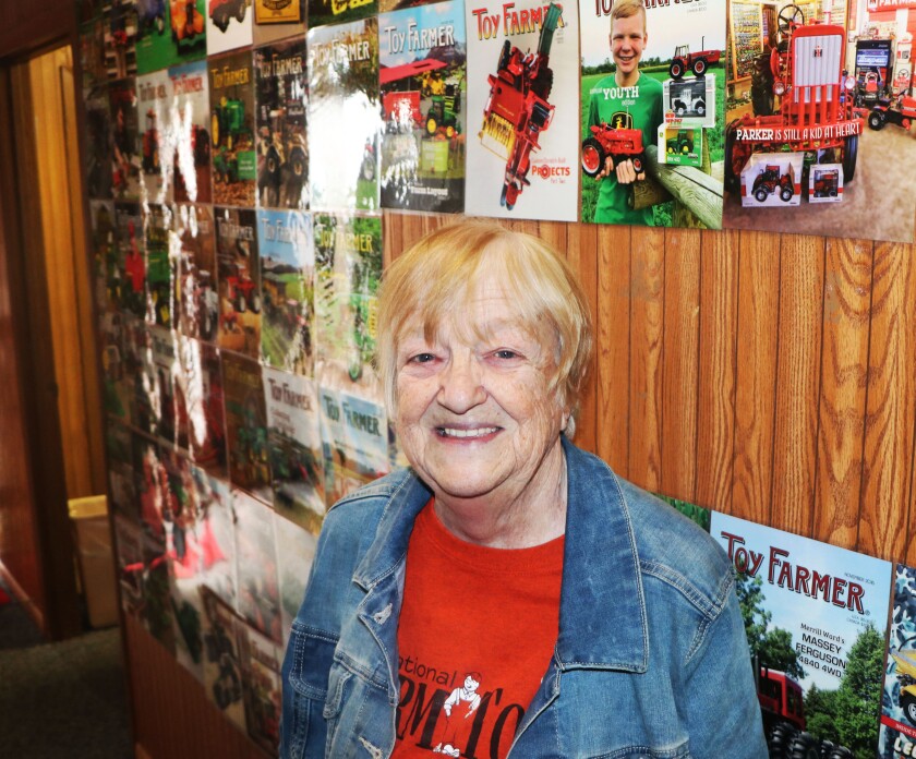 A smiling woman in a blue jean jacket and red t-shirt emblazoned with a National Farm Toy Show logo, smiles.
