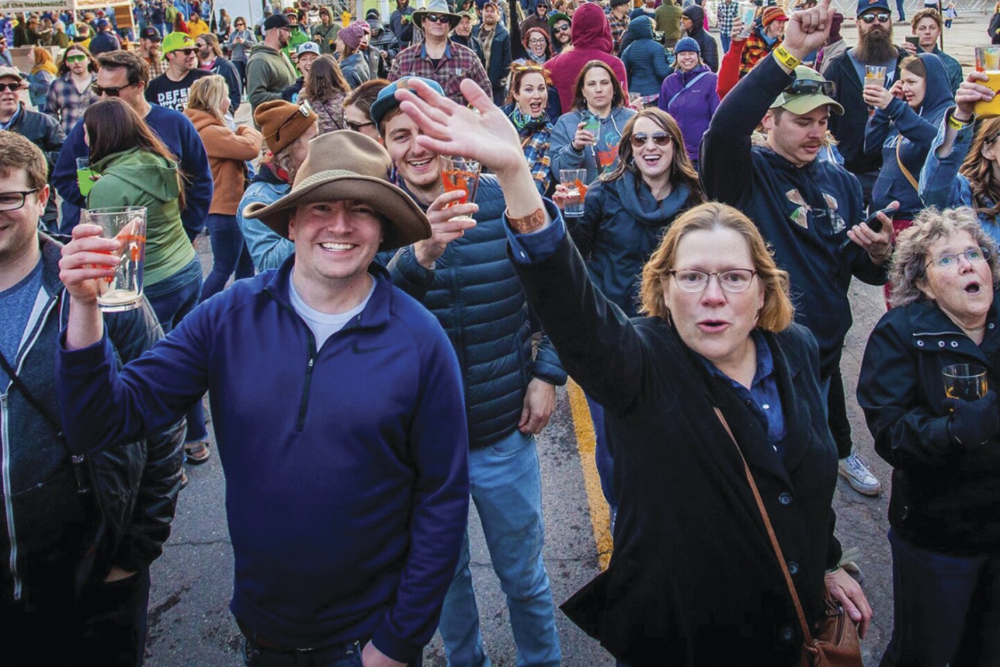 A large group of light-skinned people smile and wave to a camera. One hoists a nearly-empty glass of beer, another waves.