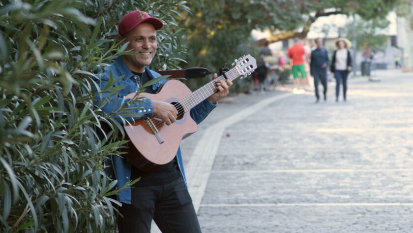 A smiling man plays a guitar in front of olive trees.
