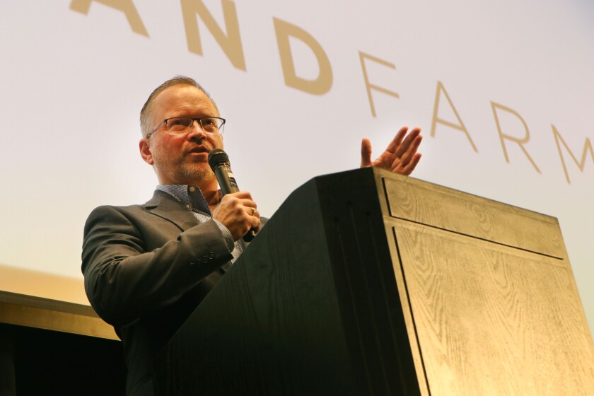 A man speaks behind a podium, gesturing, with a banner reading Grand Farm on a screen behind him.