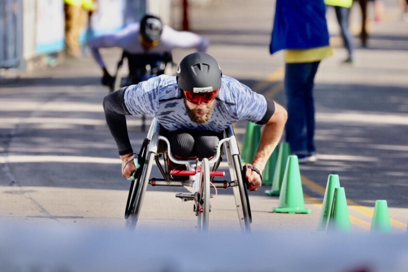 Two marathon wheelchair racers, one in front and one behind, approach a finish line