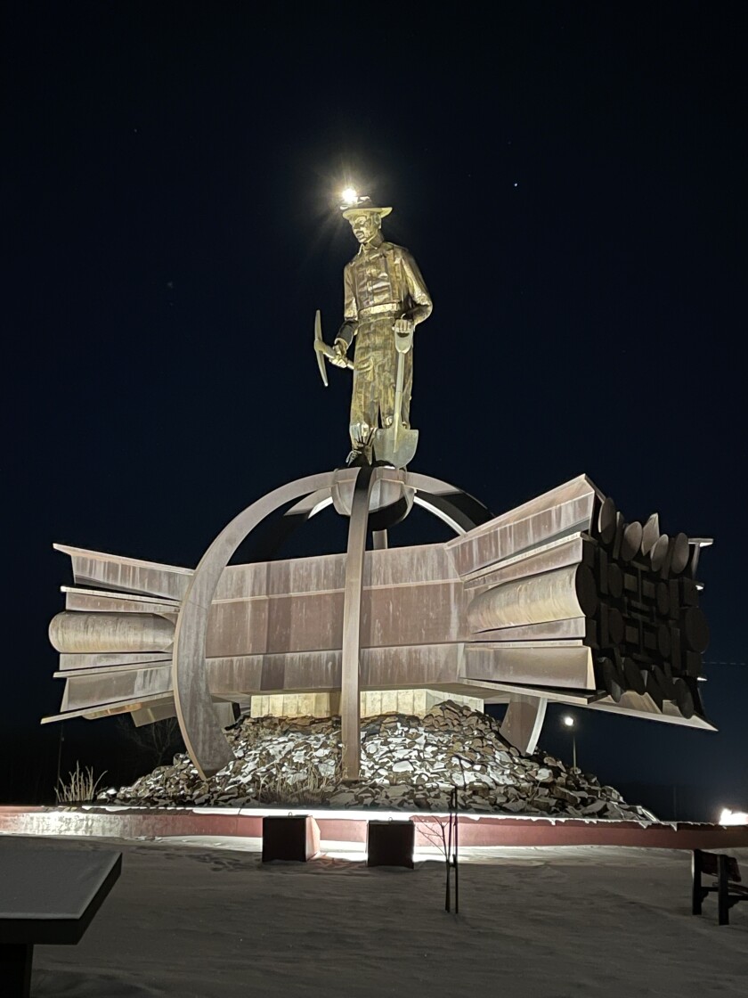 A tall statue of a miner standing on a bundle of iron beams is seen at night, the figure's head lamp illuminated and the statue underlit.