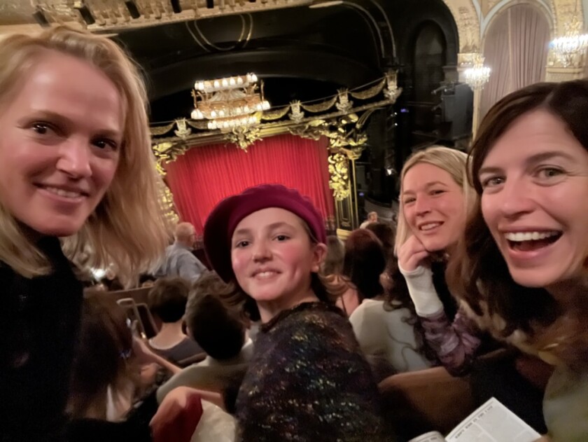 Three light-skinned women and a light-skinned girl pose for a selfie in front of red curtain and stage in theater.