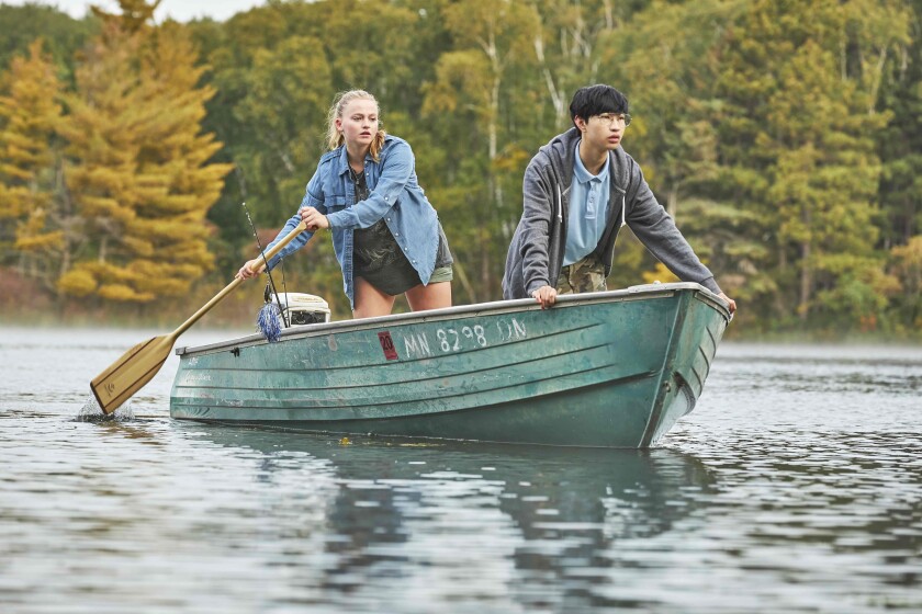 Adolescent girl and boy standing in rowboat that the girl is paddling across a lake as the boy casts his gaze forward.