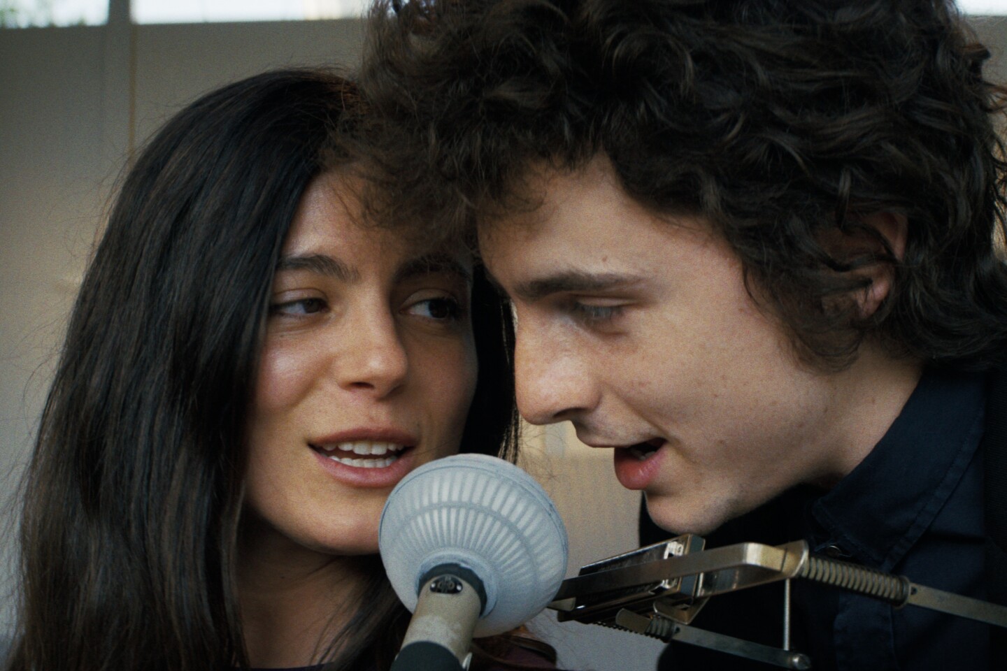 Woman in her 30s and man in his 20s sing closely together at a microphone, the man with a microphone in a rack around his neck.