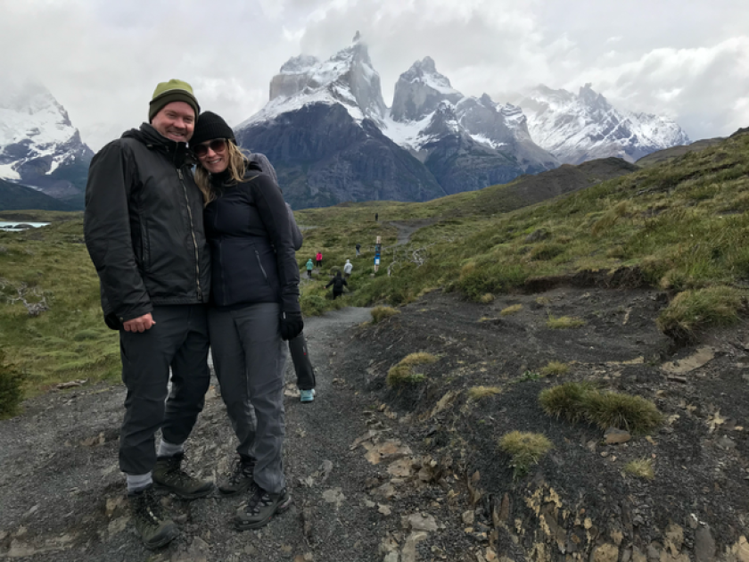 A couple in winter gear smile in front of a lush, green landscape with snow-capped mountains looming over it.