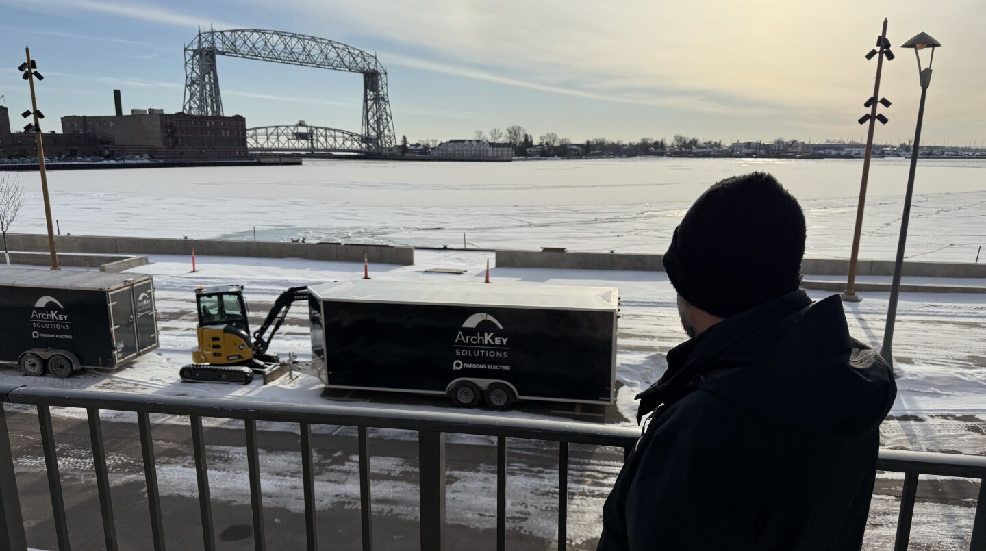 Man in black jacket and winter hat is seen from behind, looking over lake harbor with lift bridge in background. Railing stands behind him.