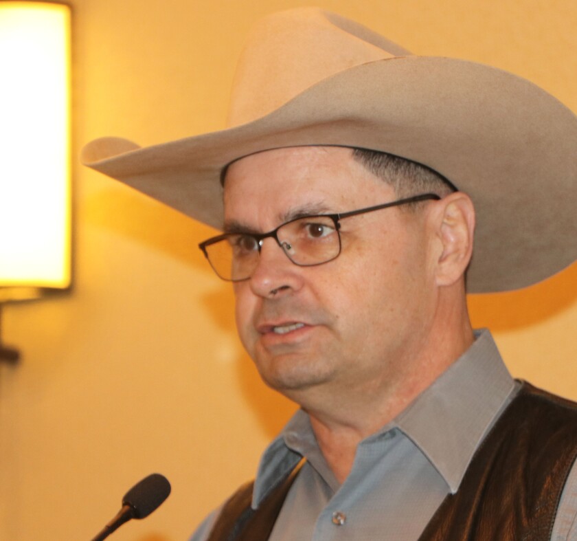 A man with eyeglasses and a cattleman's cowboy hat speaks with a microphone in a hotel conference room.