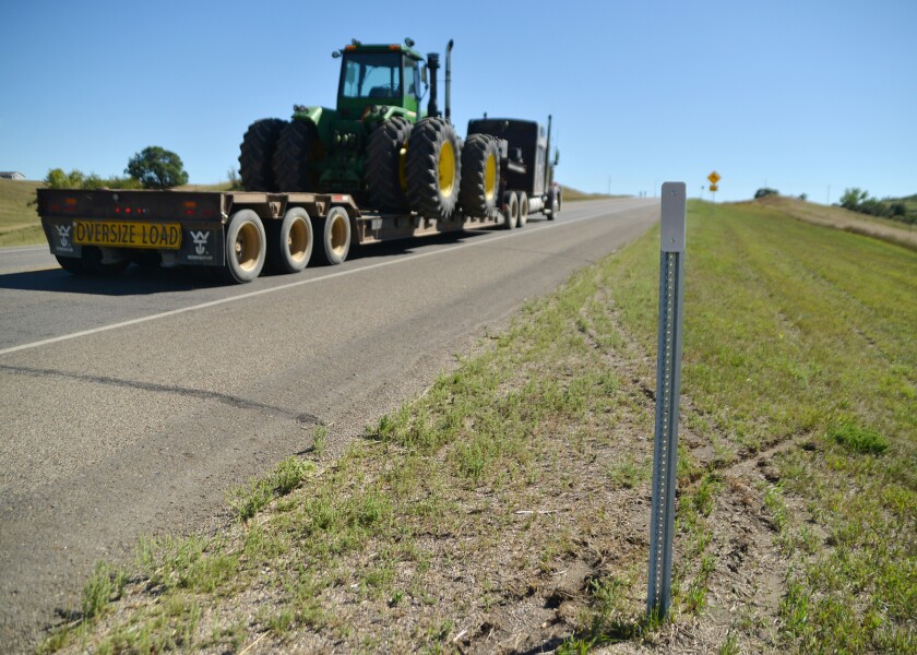 A semi hauls farm machinery down a two-lane highway, with a reflective metal post in the foreground
