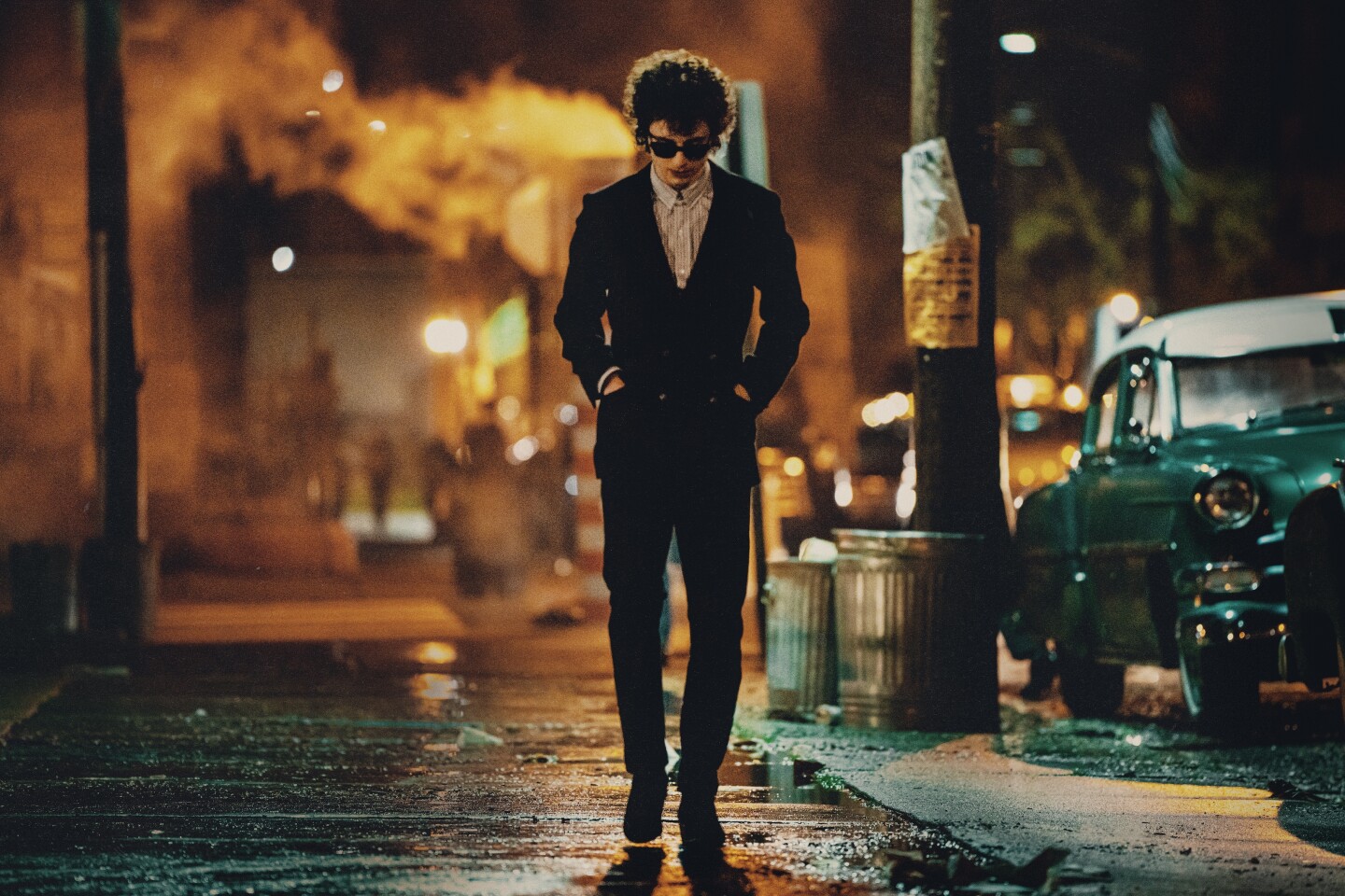 White man in his 20s, wearing a trim dark suit, walks down the a city sidewalk at night. He wears sunglasses and has his hands in his pockets.