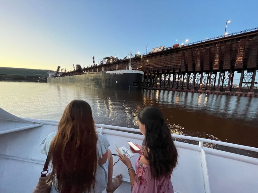 A view from the deck of the Vista Star. In the foreground, two young women are seen from behind; one holds a smartphone and a notebook featuring an illustration of a musician. In the background, the American Integrity is docked at a tall ore dock.