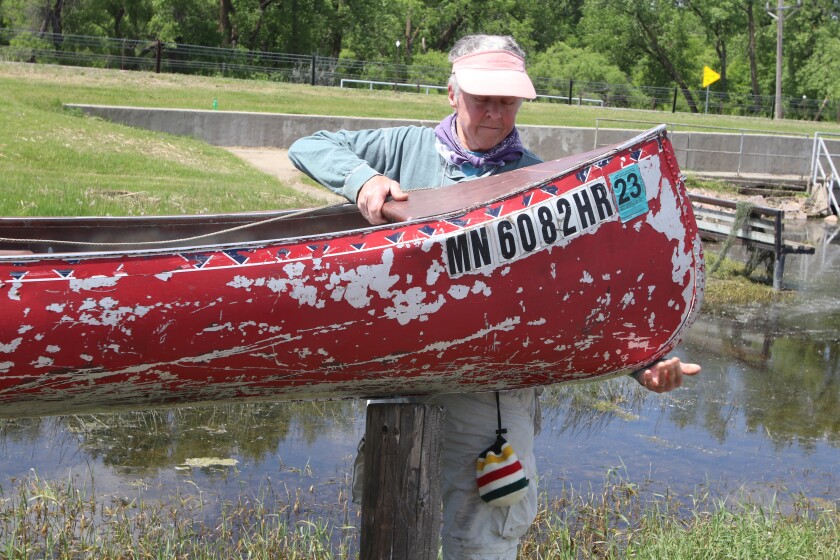 A grey-haired woman wearing a visor lifts a canoe towards a lake.
