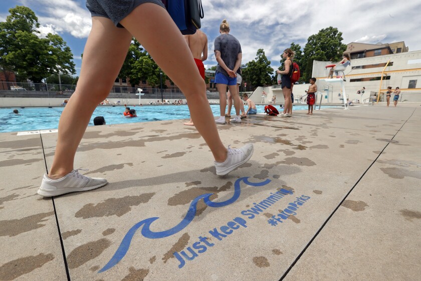 A person strides past a painted sign on concrete that says "Just Keep Swimming" with a pool in the background.