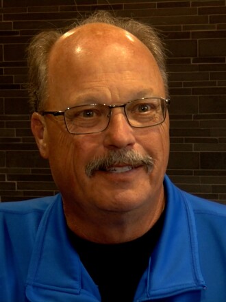 A man with a blue shirt and glasses looks at the camera. There is a brick wall behind him.
