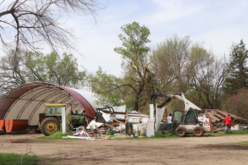 A John Deere tractor and a Bobcat skid-steer loader stand poised to dismember the remains of a wind-destroyed garage, flanked by a hoop barn on a farm.