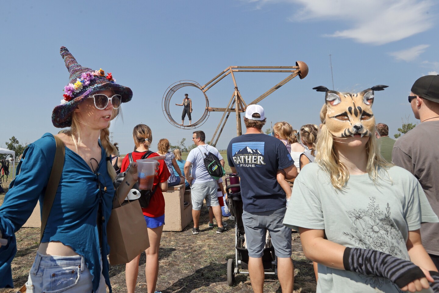 Two people, one in a bobcat mask, at a Renaissance fair with a large wooden wheel contraption in the background.