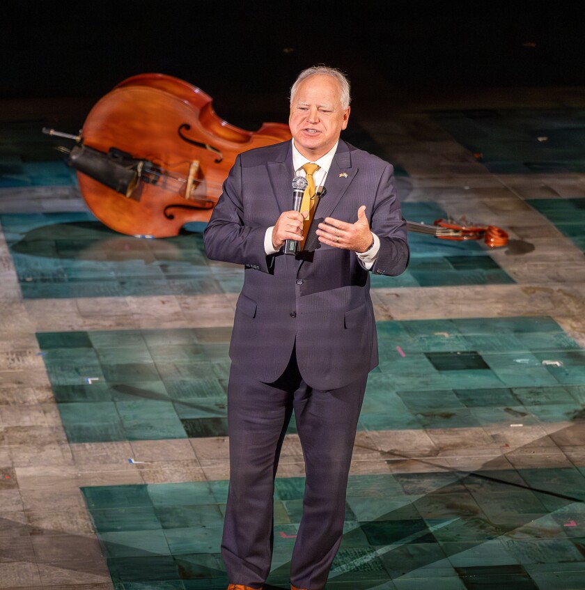 White man of advanced middle age stands on a tiled stage in front of a double-bass lying on its side. He wears a suit and holds a microphone.