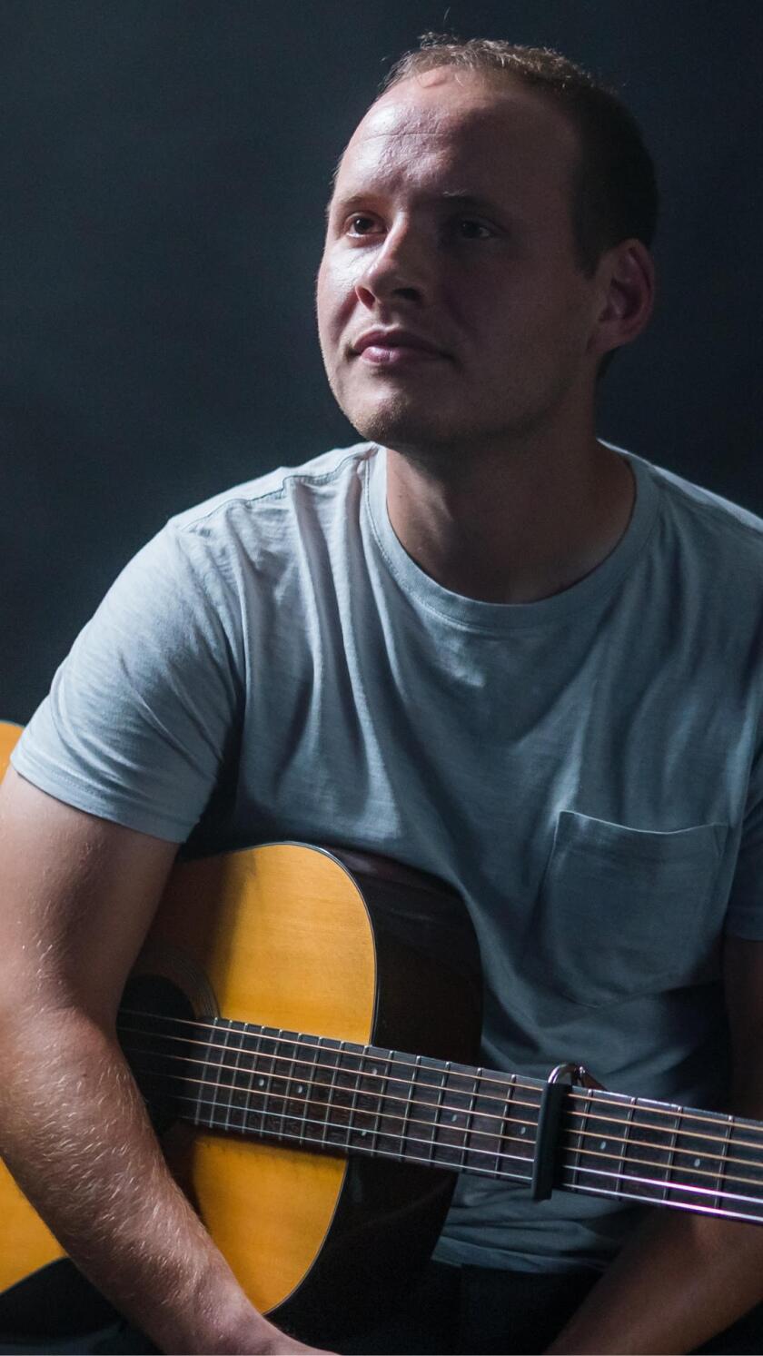 Jared Justin, a young white man, sits for a portrait holding an acoustic guitar and looking offscreen to the left.