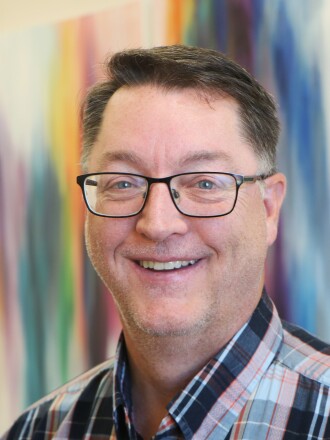 A smiling man with glasses is flanked by a colorful impressionistic art piece on the wall.