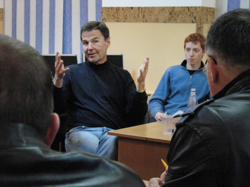 Howard Dahl, dressed in a black shirt and blue jeans, flanked by an engineer-employee, gestures joyfully as he talks about sugar beet equipment with a group of Ukrainian farmers.