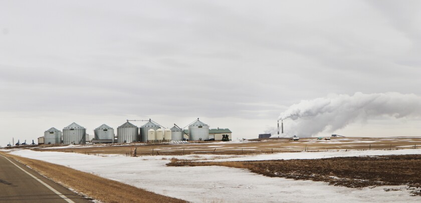 A large, modern farmstead, and its grain bins, stand at the top of a hill in early March snow remnants. Behind it several miles are the smokestacks of a coal-fired electrical power plant, which is next to an ethanol plant.