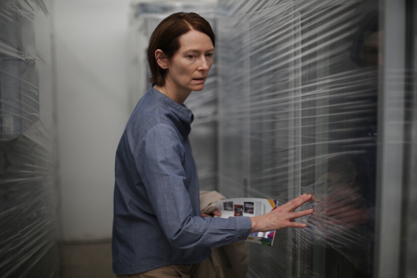 A white woman of middle age stands between two walls lined with sheets of plastic, holding a brochure and looking pensive.