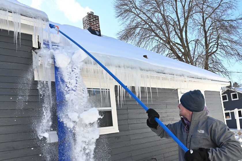 A man pulls snow off the roof with a roof rake.