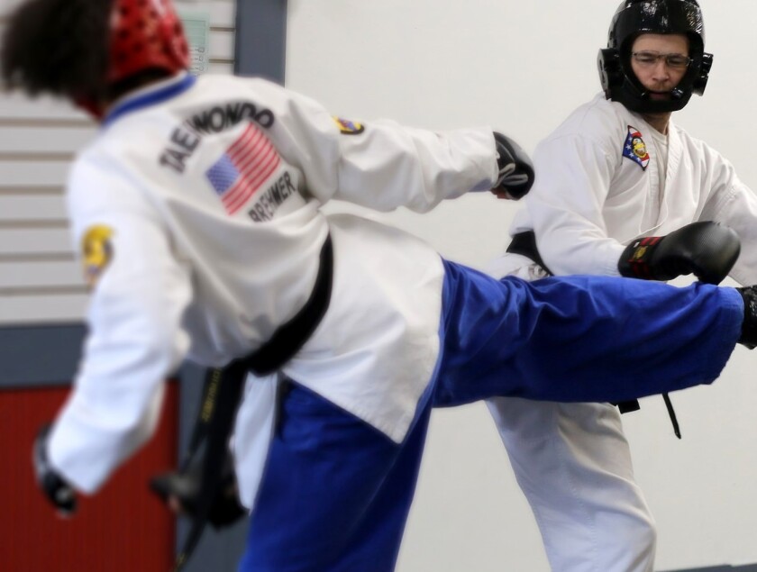 Two athletes spar in taekwondo, wearing blue pants, white shirts and padded helmets. Athlete in foreground is kicking, while opponent attempts to dodge.