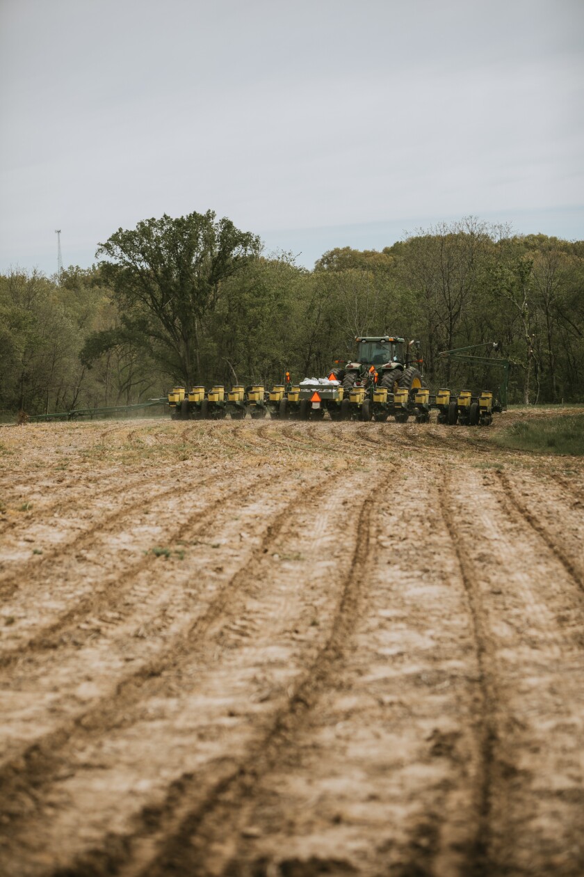 The foreground shows the light marks left in the soil from a no-till drill planting corn. The John Deere tractor and the planter are visible in the background, traveling down a field. The far background of the photo features a grove of trees.
