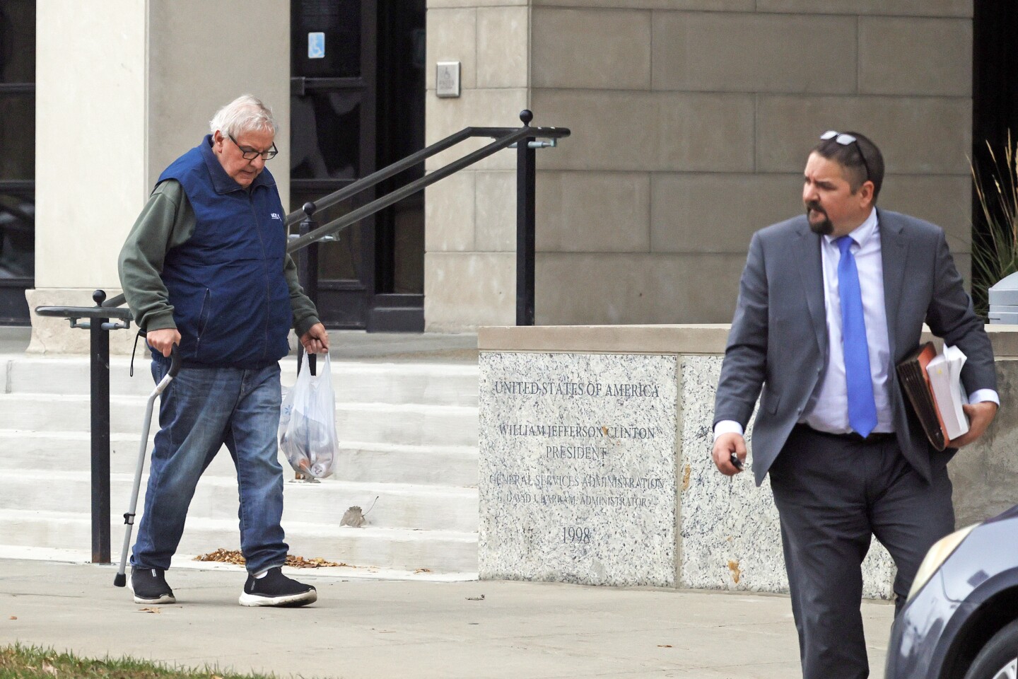 In a blue vest and green long-sleeve shirt, Ray Holmberg uses a cane, trailing behind a man in a suit.