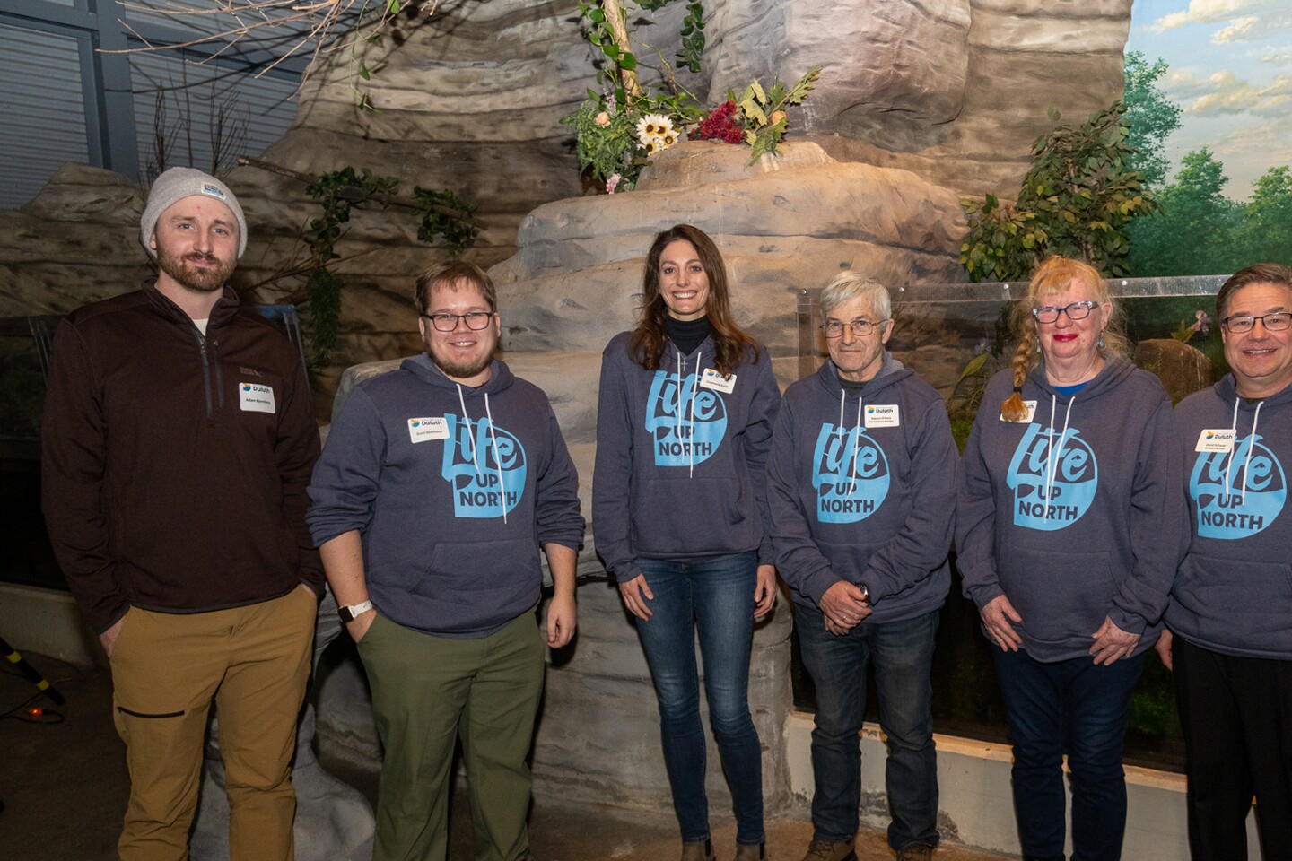 Six light-skinned people stand in front of a simulated rocky outcropping with adjacent aquarium tank. Five people on right wear blue hoodies with insignia reading "life up north."