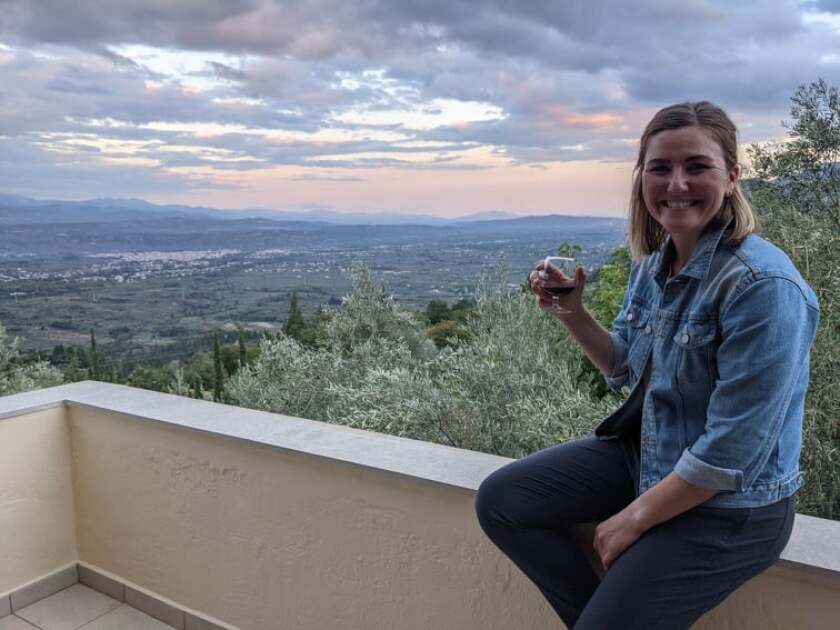 A smiling woman with a glass of wine sits on a low wall.