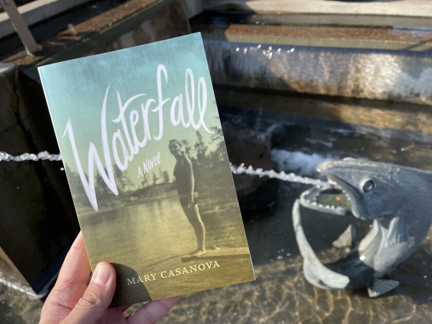 Hand holding paperback book: Mary Casanova, "Waterfall." In the background, a fountain is visible with water shooting out of a fish's mouth and a nearby artificial waterfall.