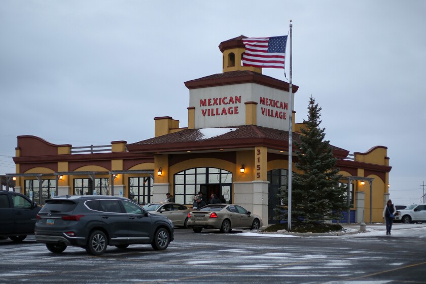 A one-story building with a two-story tower on one corner that reads "Mexican Village." A U.S. flag flies in front of the building under a gray sky. Several cars are parked in the parking lot that surrounds the building.