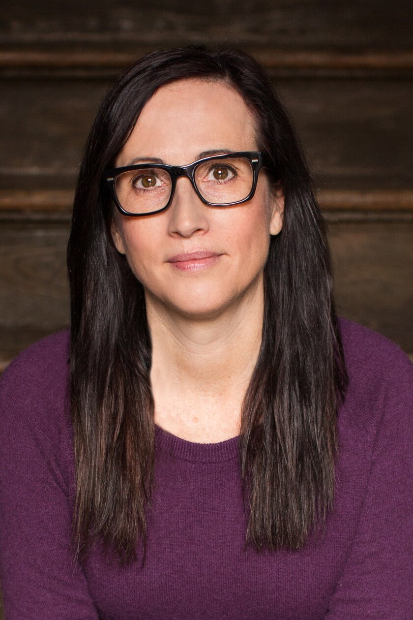A white-presenting woman sits for a headshot. She has long brown hair, dark-framed eyeglasses, and a purple sweater.