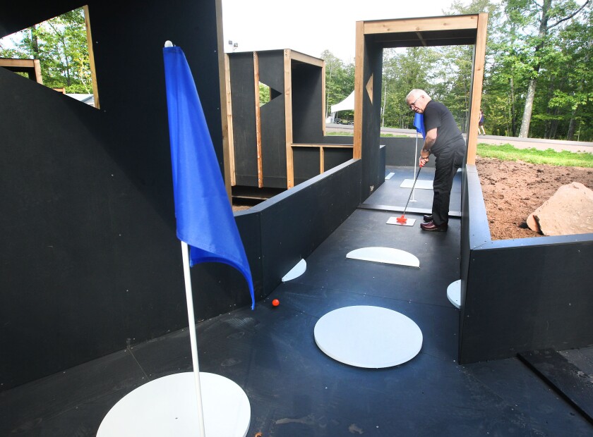 White man in his 70s prepares to putt a ball in a miniature golf course where the surface is completely black, surrounded by black walls that rise to the top of his legs.